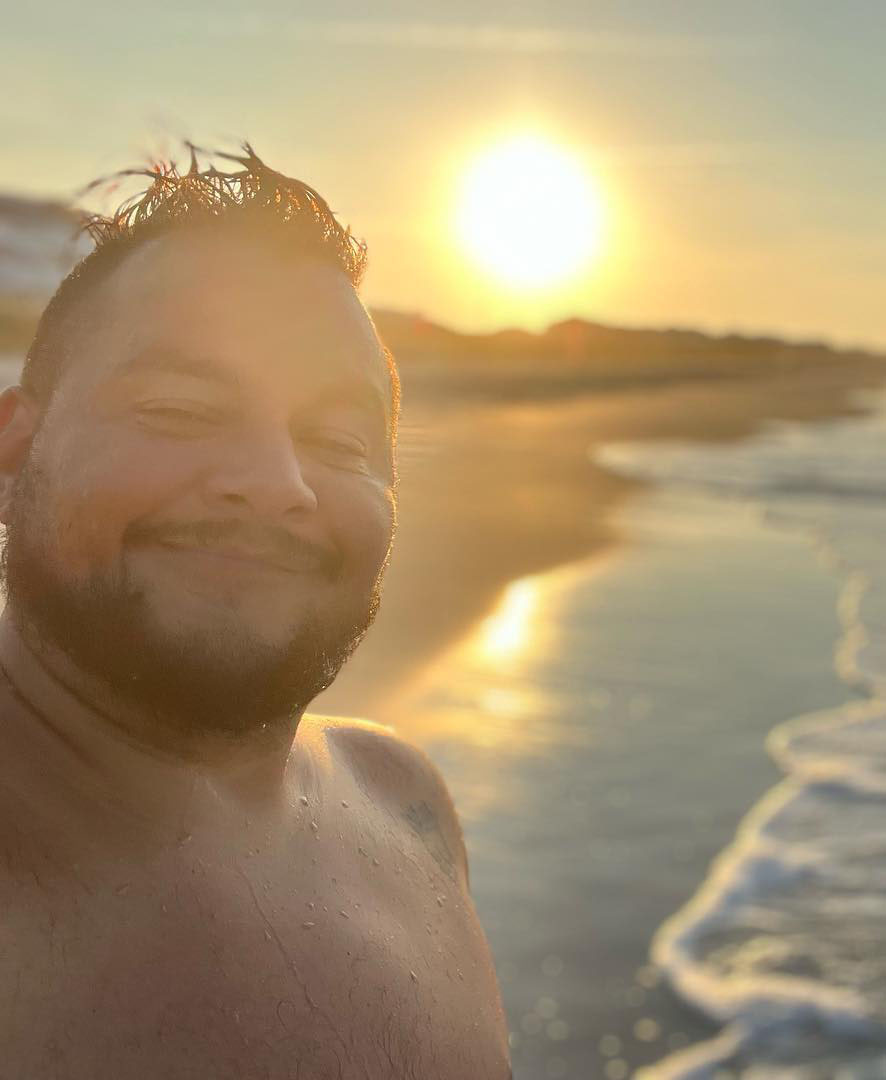 Jack Knoxville is a medium brown skinned trans man shown smiling on the beach