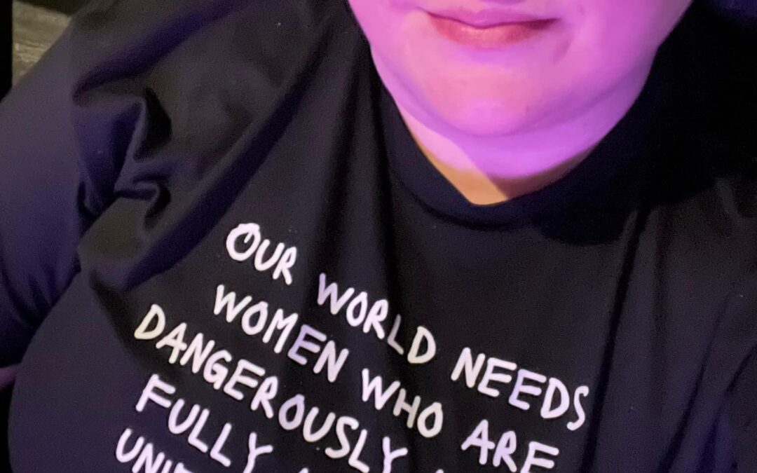 Heather Knoxville is a white woman wearing multi colored reading glasses and a black t-shirt which reads "Our world needs women who are dangerously awake, fully alive and unified in heart and purpose."