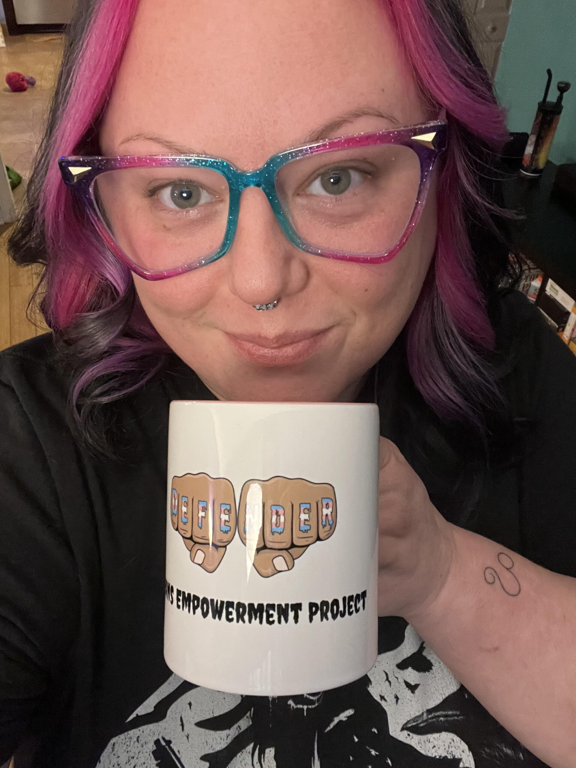 Heather Knoxville is a white woman with shoulder length pink hair, smiling while holding up a  white coffee mug, branded with Trans Empowerment Project messaging. 