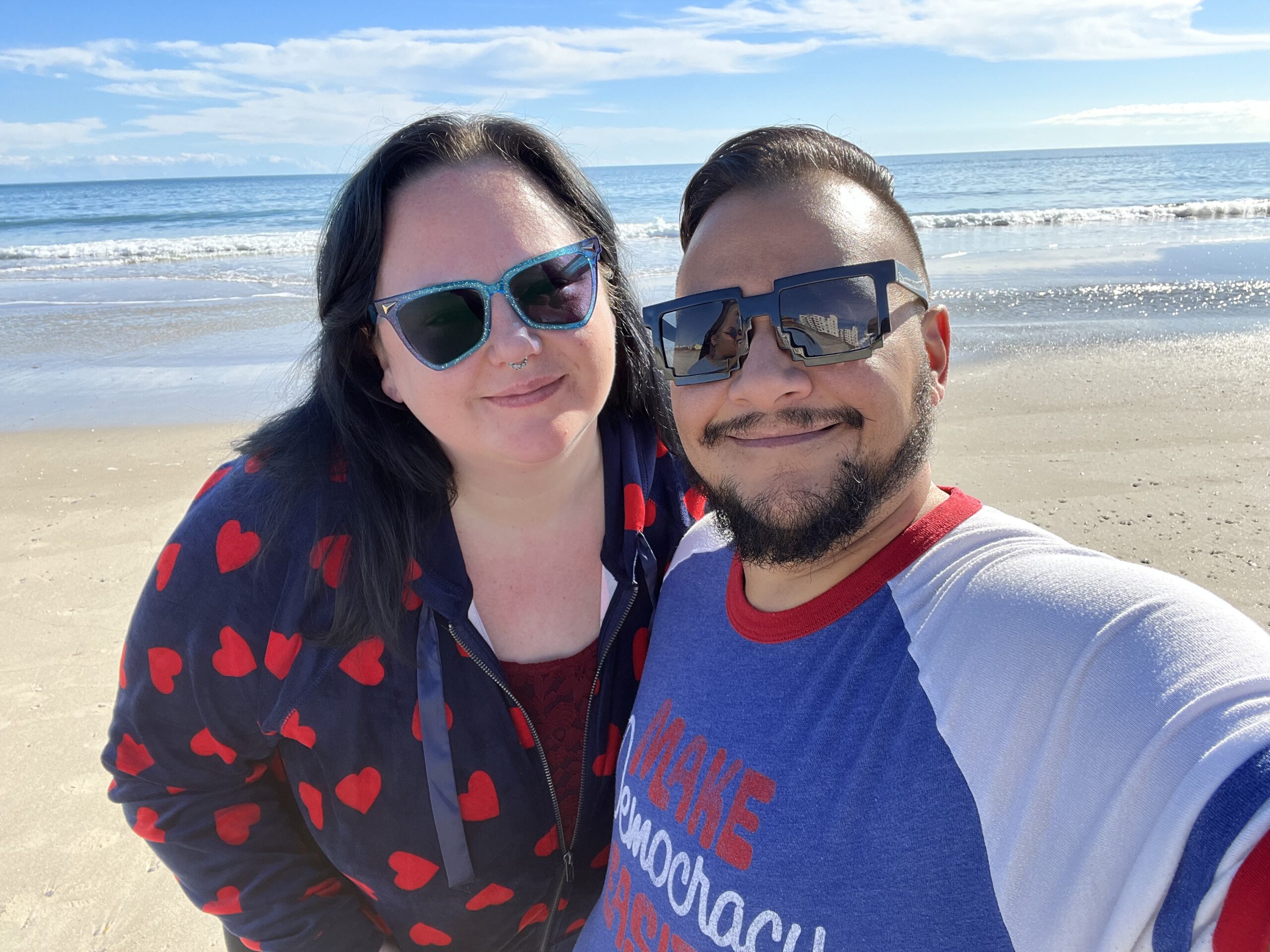 Jack and Heather Knoxville are shown together smiling. Jack is a medium brown skinned man with medium slick backed hair and dark glasses. He has a beard and mustache. Heather has dark brown hair styled into two small buns on either side. She is wearing a dark Tshirt under a purple and black jumper.