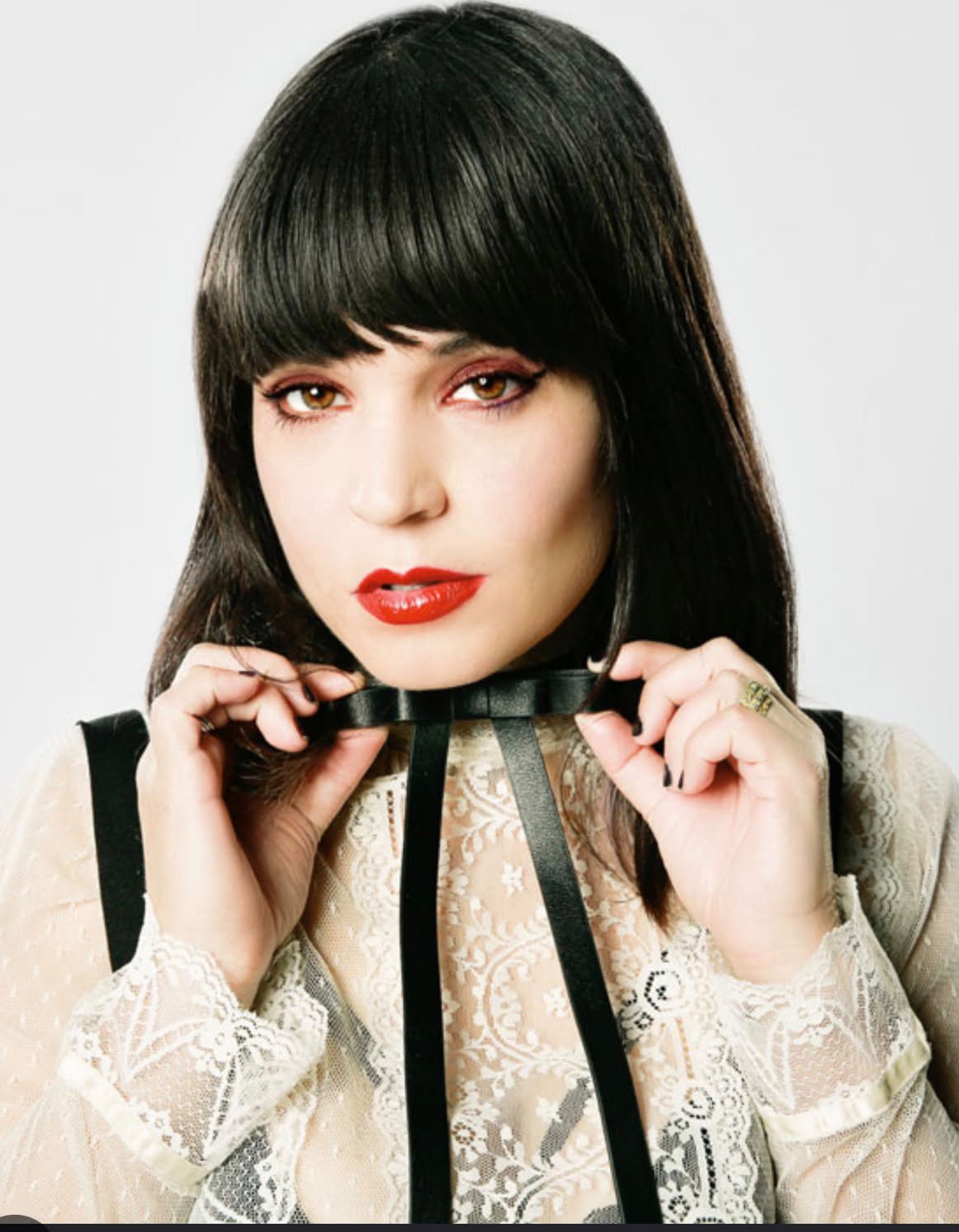In a world of Joannes be a Jessicka campaign graphic shows Jessicka Addams a white woman with long black hair with cropped bangs. Wearing an off white blouse with a thin black bow tie in front of a large yellow heart